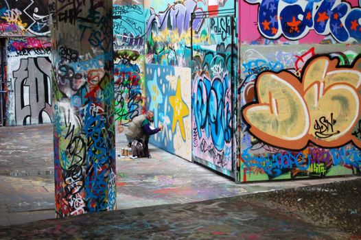 Artiste en train de peindre une fresque colorée sur un mur recouvert de graffitis dans un espace urbain dynamique.